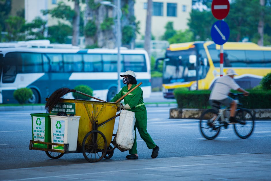 A person dressed in an orange cleaning jumpsuit, wearing white gloves and a protective face mask with goggles, stands outdoors against a grey concrete wall. They are holding two large green garbage ba